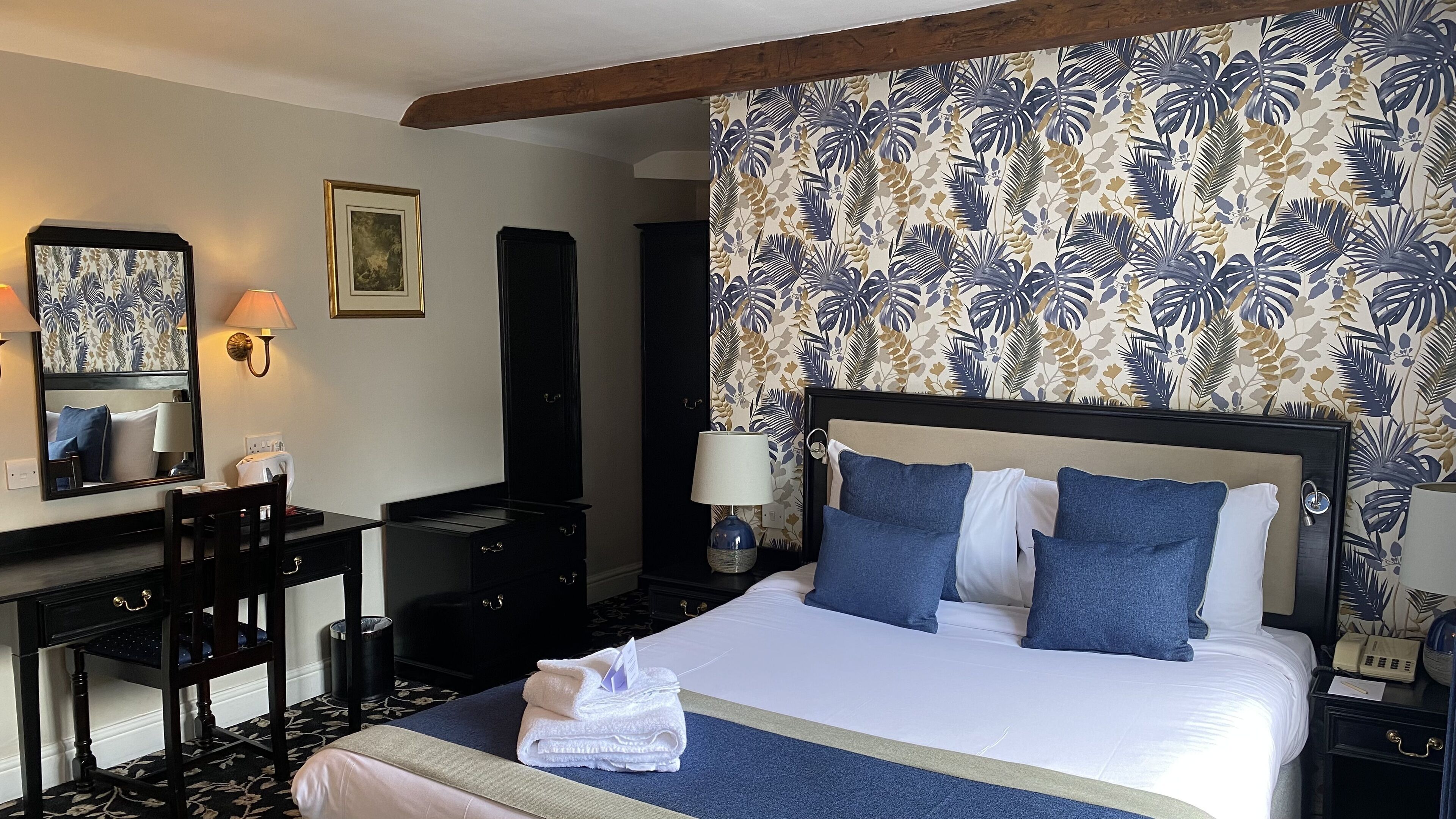 A hotel room with a large bed, a desk with a chair, a dresser, a nightstand with a lamp, and a wall with a tropical wallpaper pattern.