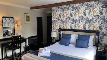 A hotel room with a large bed, a desk with a chair, a dresser, a nightstand with a lamp, and a wall with a tropical wallpaper pattern.