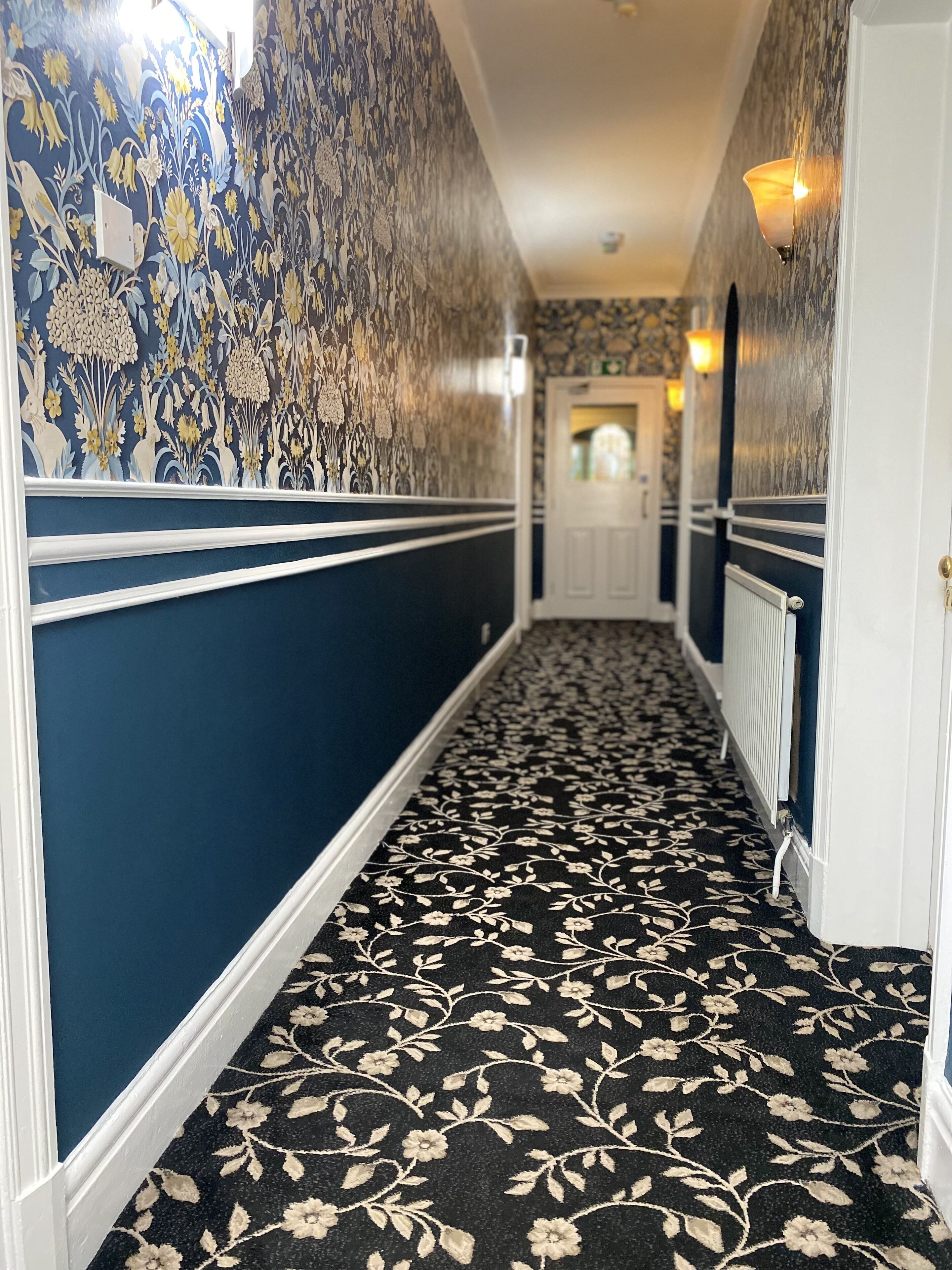 A hallway with patterned wallpaper and carpet, white trim, and wall sconces.