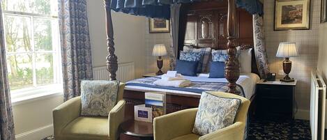 A four-poster canopy bed with a blue bedspread, two armchairs with patterned cushions, a bedside table with a lamp, and a framed picture on the wall.