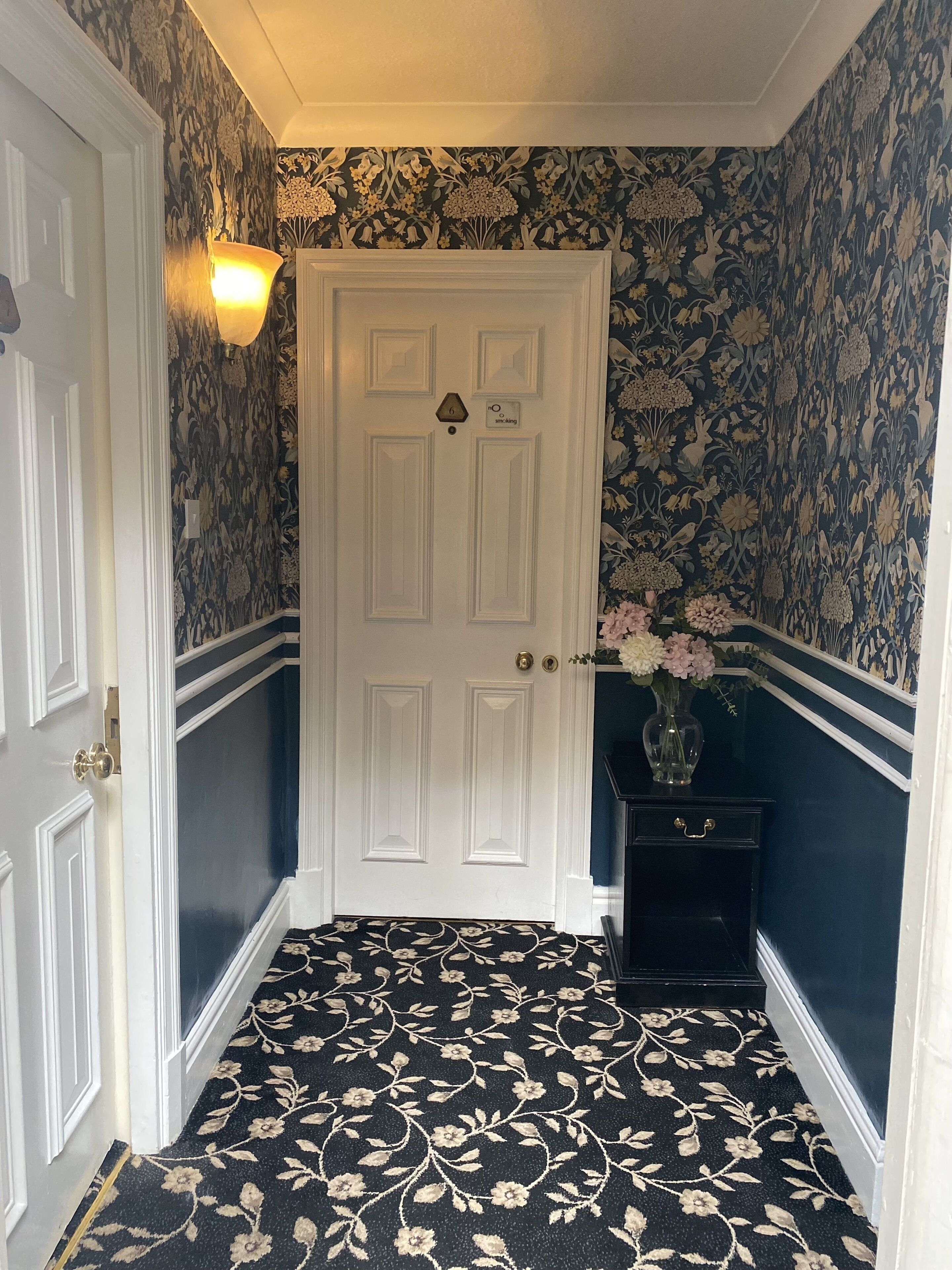 A hallway with a white door, floral wallpaper, a black and white patterned carpet, and a vase with flowers on a black side table.