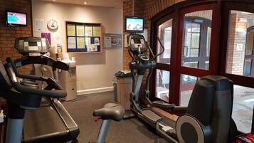 A gym with cardio machines, a digital clock, and a TV screen displaying a tennis match.