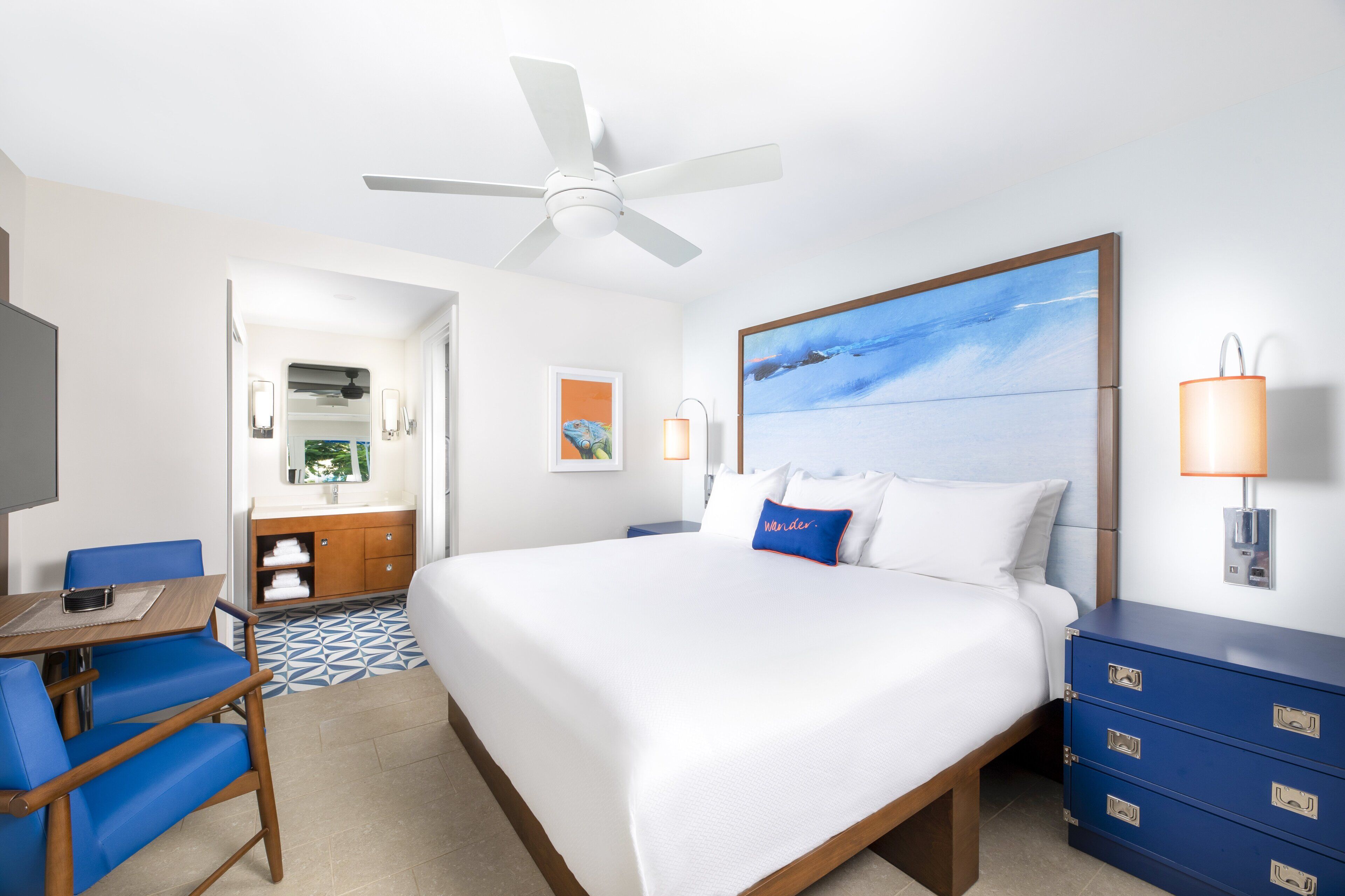 A hotel room with a bed, a desk with a chair, a dresser, a ceiling fan, and a painting on the wall.