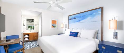 A hotel room with a bed, a desk with a chair, a dresser, a ceiling fan, and a painting on the wall.