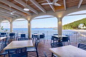 An outdoor dining area with tables and chairs, a wooden ceiling, and a view of the sea.