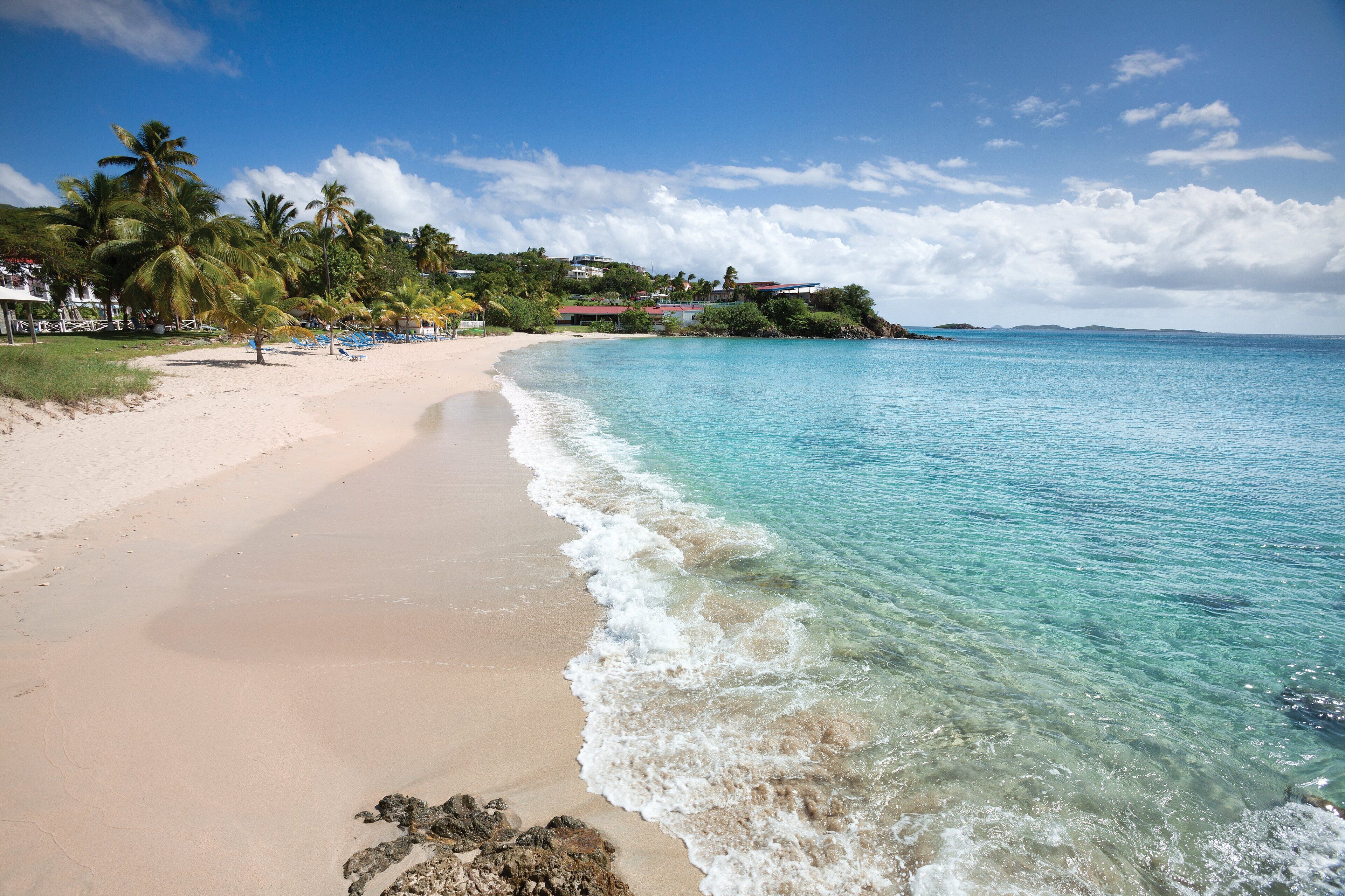 Ein Strand mit klarem türkisblauem Wasser, sandigem Ufer, Palmen und Häusern im Hintergrund.