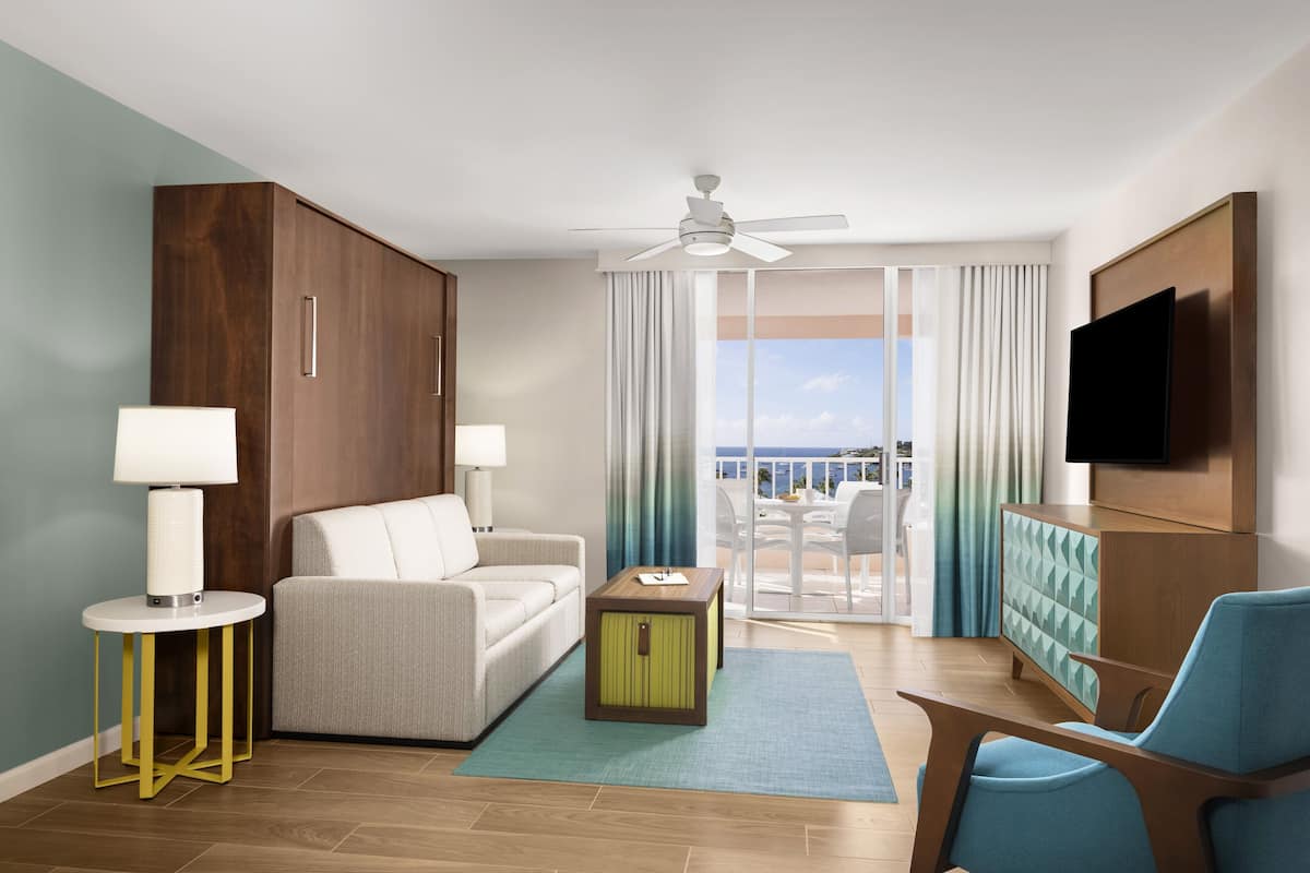 Una habitación de hotel moderna con sofá, una mesita, un armario, televisor y balcón con vistas.