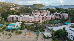 Ein Resort mit mehreren Gebäuden, einem Strandabschnitt mit Sonnenschirmen und Blick auf das Meer und die Hügel.