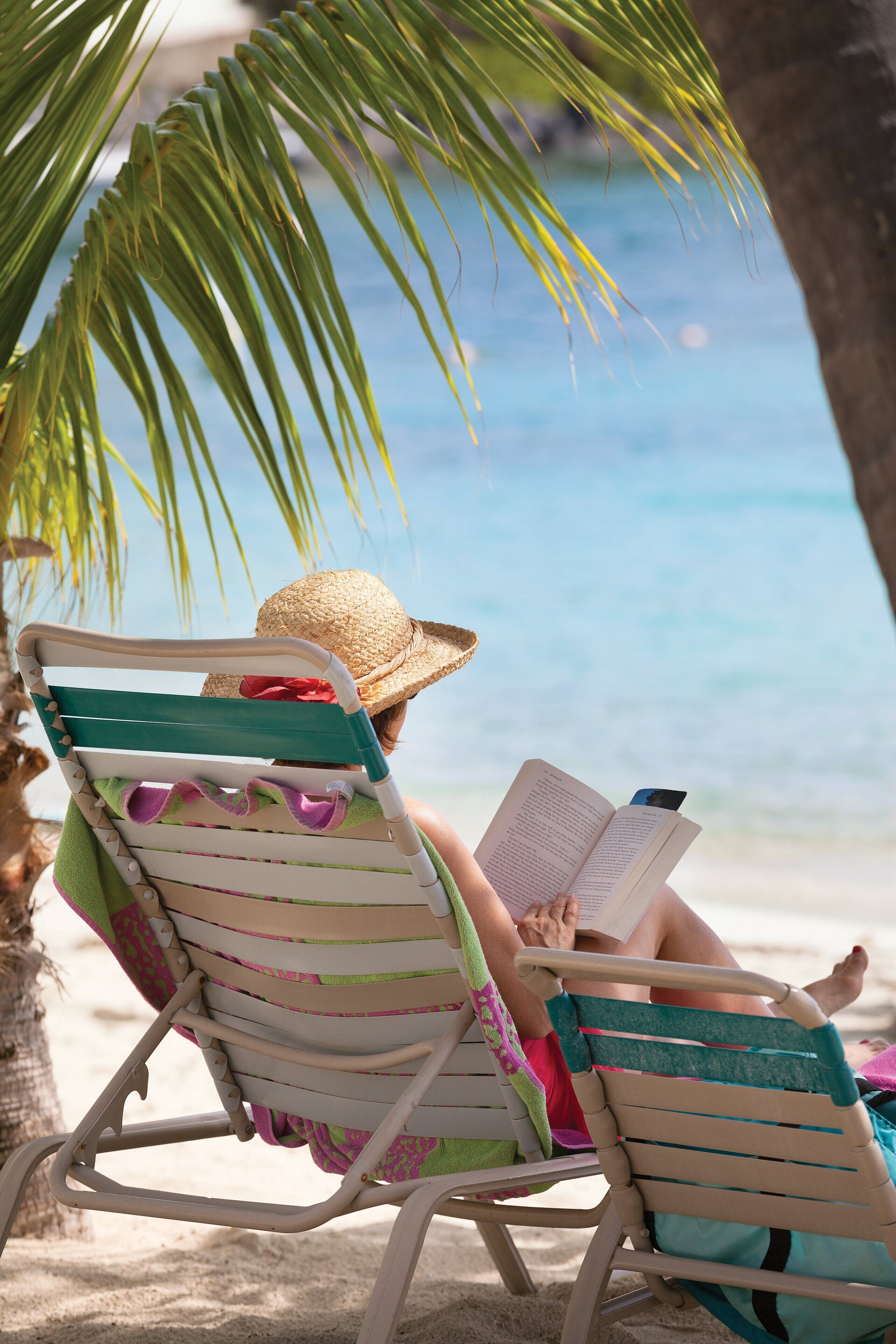 A person reading a book in a beach chair with a straw hat, under a palm tree, by the ocean.