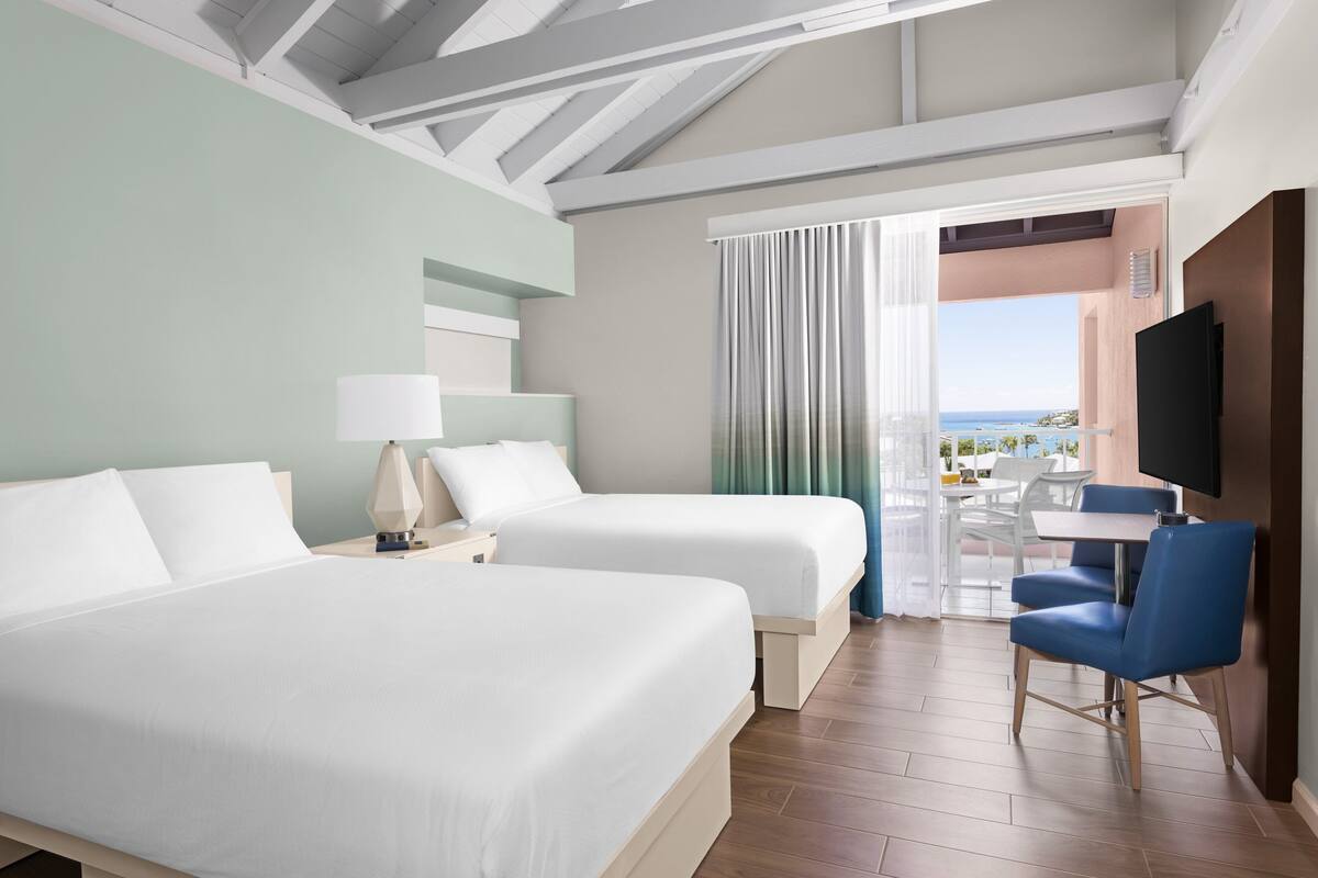 Habitación de hotel con dos camas, televisión, escritorio con silla y vista al mar.