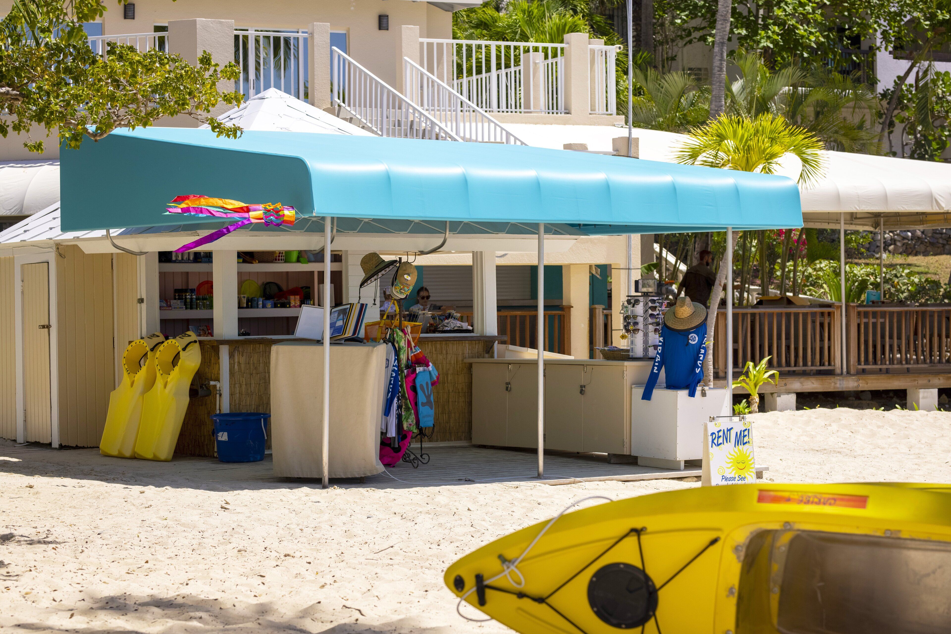 Un petit kiosque situé sur la plage, doté d’un auvent bleu, d’une enseigne indiquant les services de location et d’un kayak jaune à proximité.