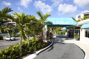 Entrada de un hotel con un toldo azul, palmeras y un camino pavimentado.