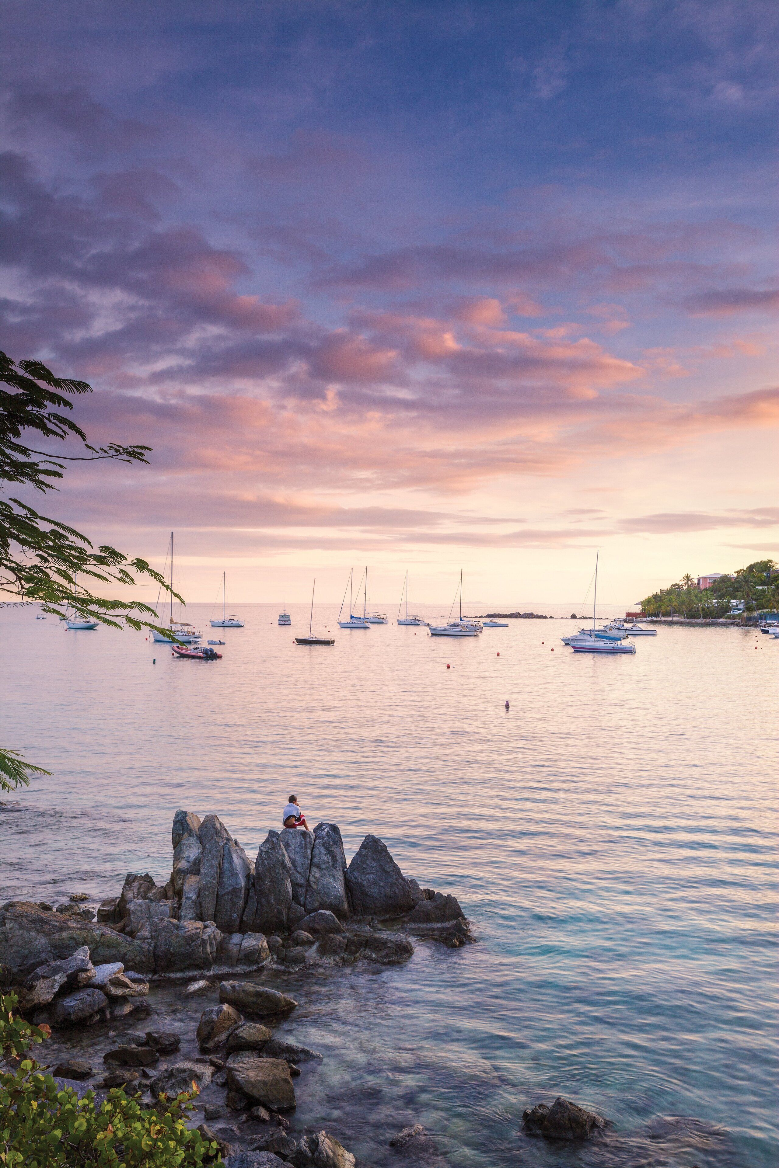 A coastal scene with boats, a rocky shoreline, and a person sitting on rocks.