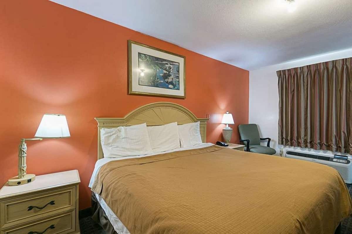 Una habitación de hotel con cama, dos lámparas de noche, una silla, un calentador y una ventana con cortinas.