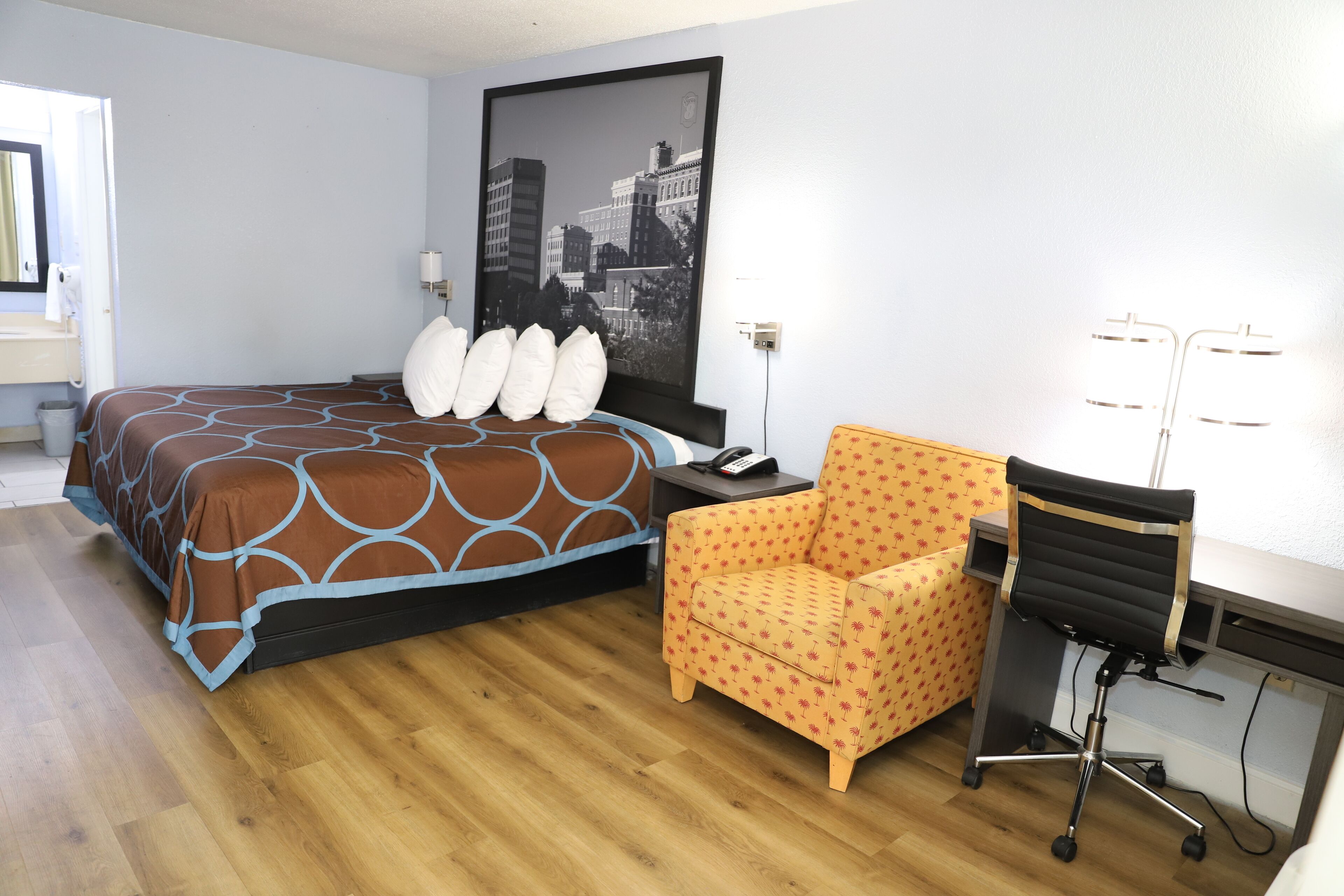 A hotel room with a bed, a desk, a chair, and a large cityscape painting on the wall.