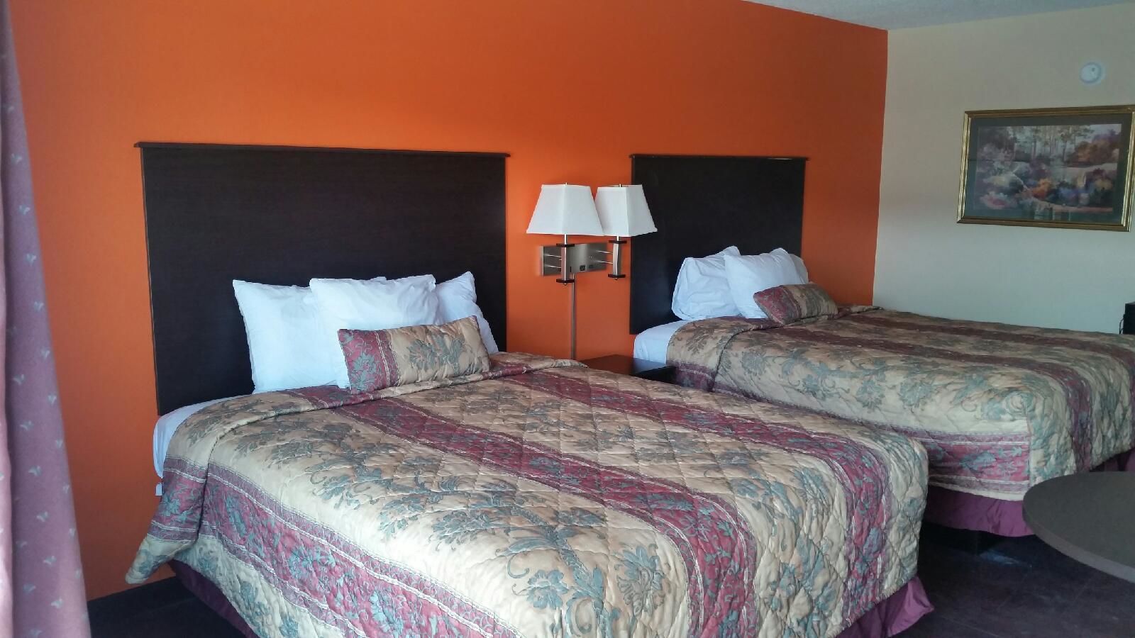 Una camera d'albergo con due letti, ognuno con una coperta a motivi e cuscini bianchi. Tra i letti, un comodino con una lampada.