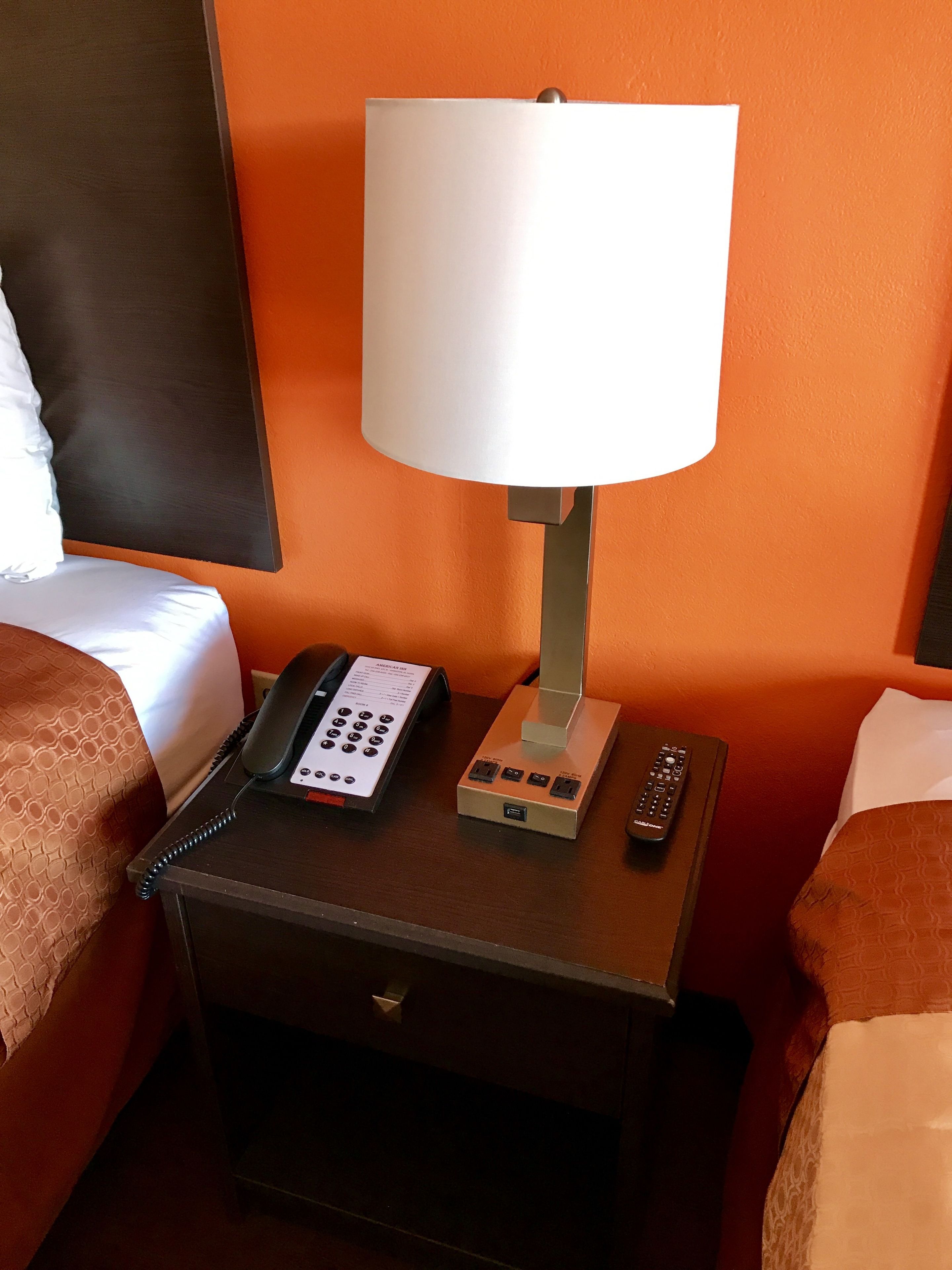 A hotel room with a bedside table featuring a lamp, a telephone, and a remote control.
