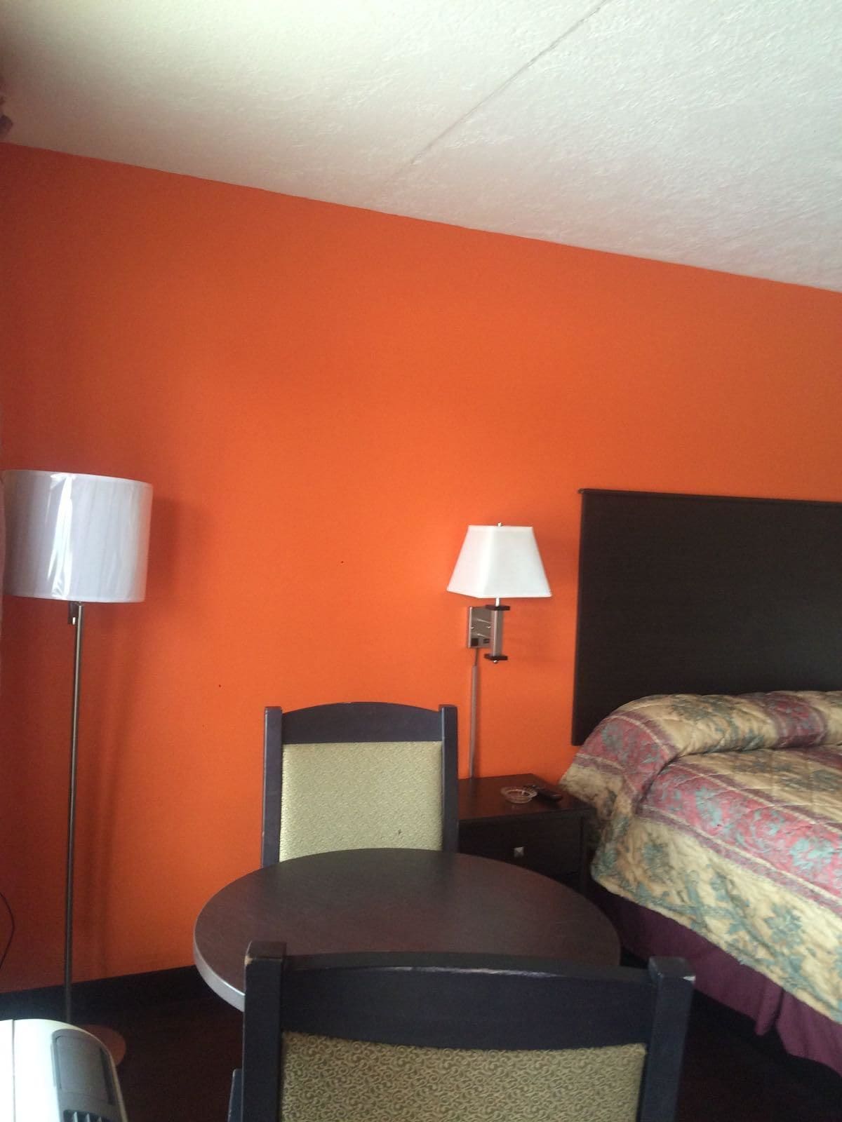 Habitación de hotel con pared naranja, cama con manta estampada, mesa redonda con una silla y dos lámparas de noche.