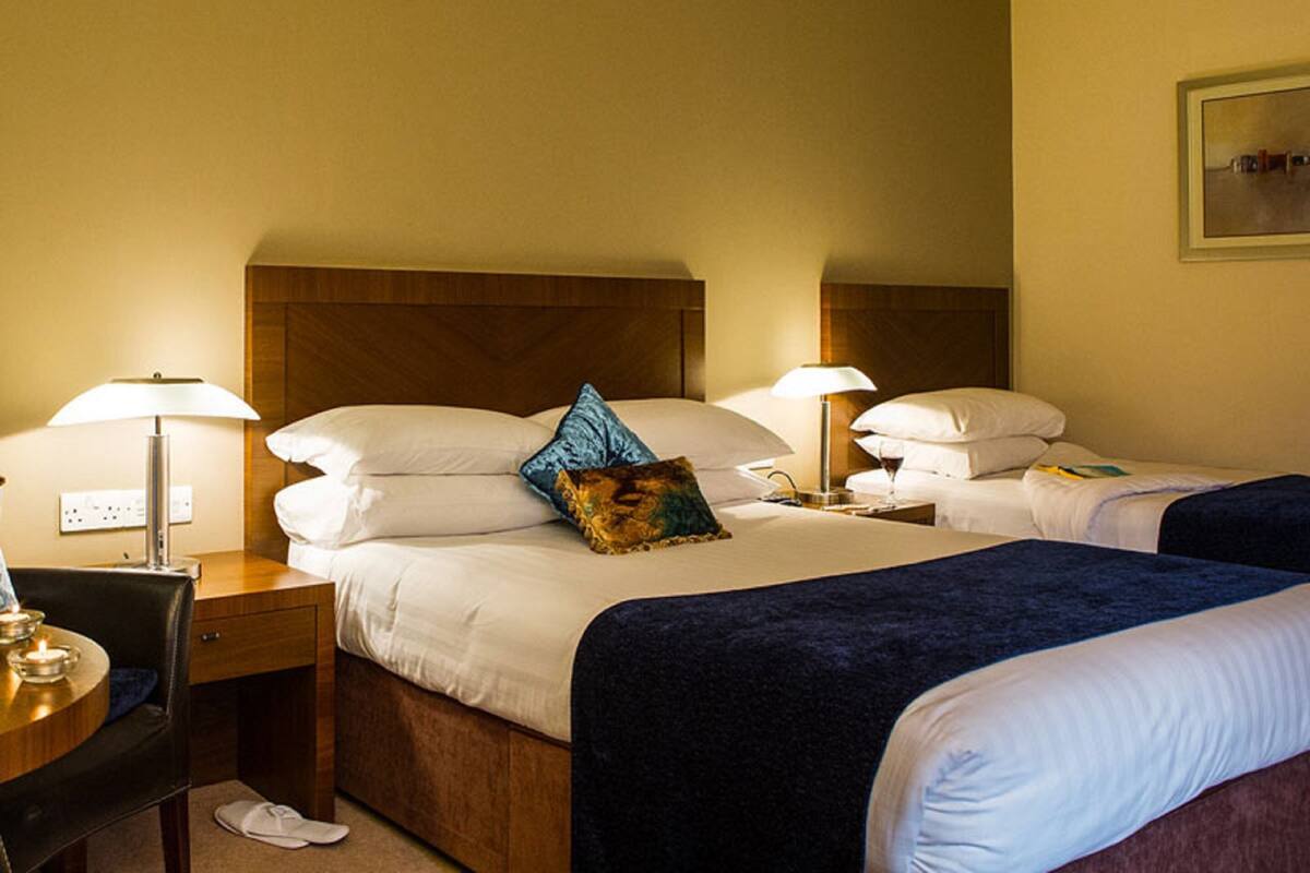 A hotel room with two beds, a desk, and a chair. There are lamps on each side of the beds.