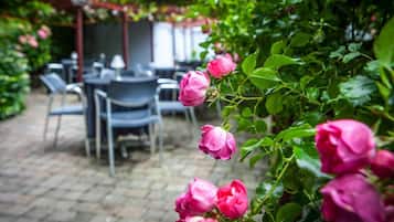 Un espace jardin agrémenté d’une terrasse en briques, de tables et de chaises, entouré d’une végétation luxuriante et de rosiers roses.