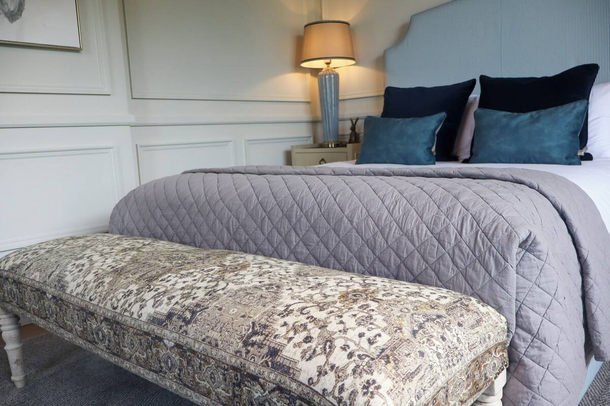 A neatly made bed with a grey quilt, a bedside lamp, and a patterned bench.
