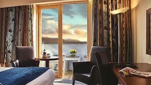 A hotel room with a bed, two chairs, a small table with a coffee pot, a vase with flowers, and a view of the sea through a large window. - Ballyroe Lodge (Tralee)