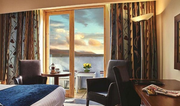 A hotel room with a bed, two chairs, a small table with a coffee pot, a vase with flowers, and a view of the sea through a large window.