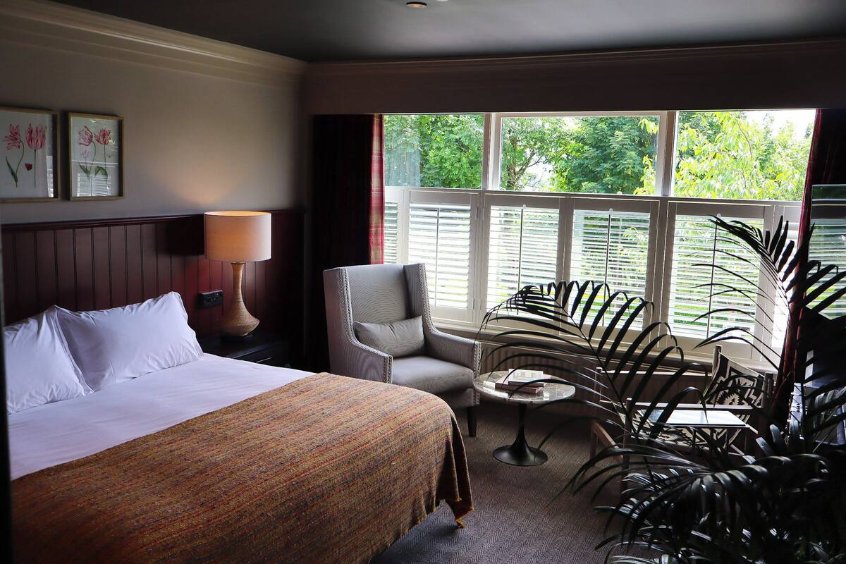 A hotel room with a bed, a chair, a table, and a window with a view of greenery.
