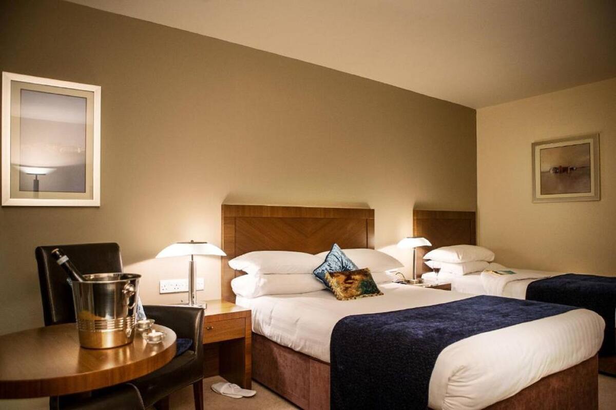 A hotel room with two beds, a wooden headboard, bedside lamps, a small table with a metal bucket, and a chair.
