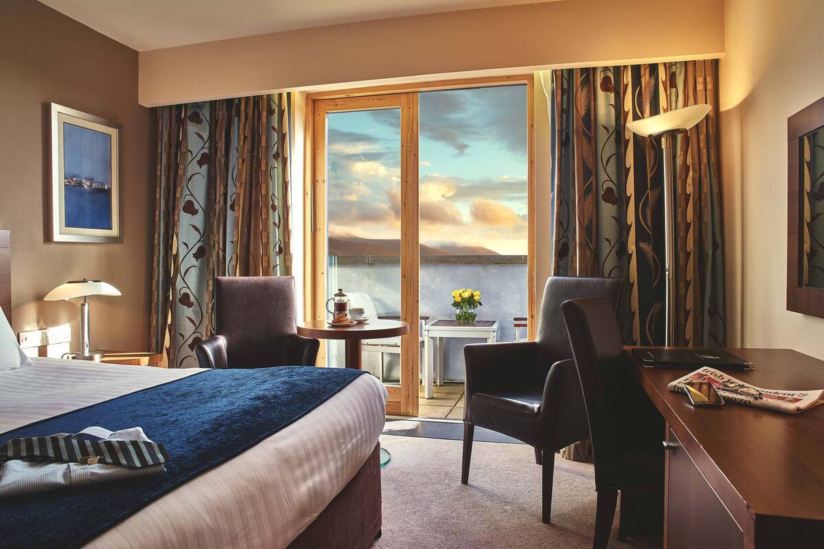 A hotel room with a bed, a desk, a chair, a table with a vase of flowers, and a view of the sky through large windows.