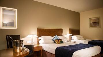 Habitación de hotel con dos camas, cabecero de madera, mesita de noche con lámpara, una silla y una mesa con un cubo.