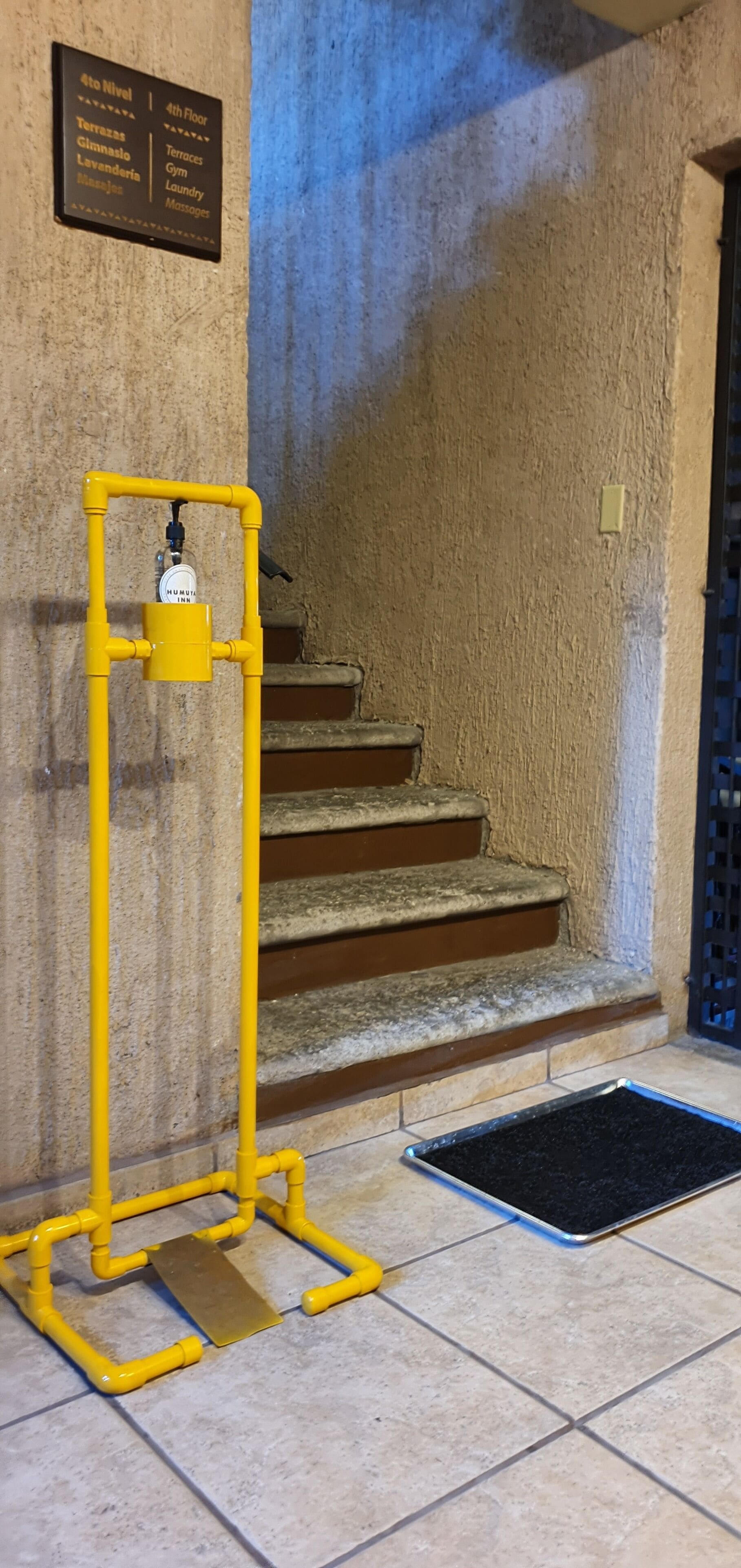 Une rambarde jaune à côté d’un escalier, un tapis noir au bas de l’escalier et un panneau indiquant les noms des pièces et les numéros des étages.