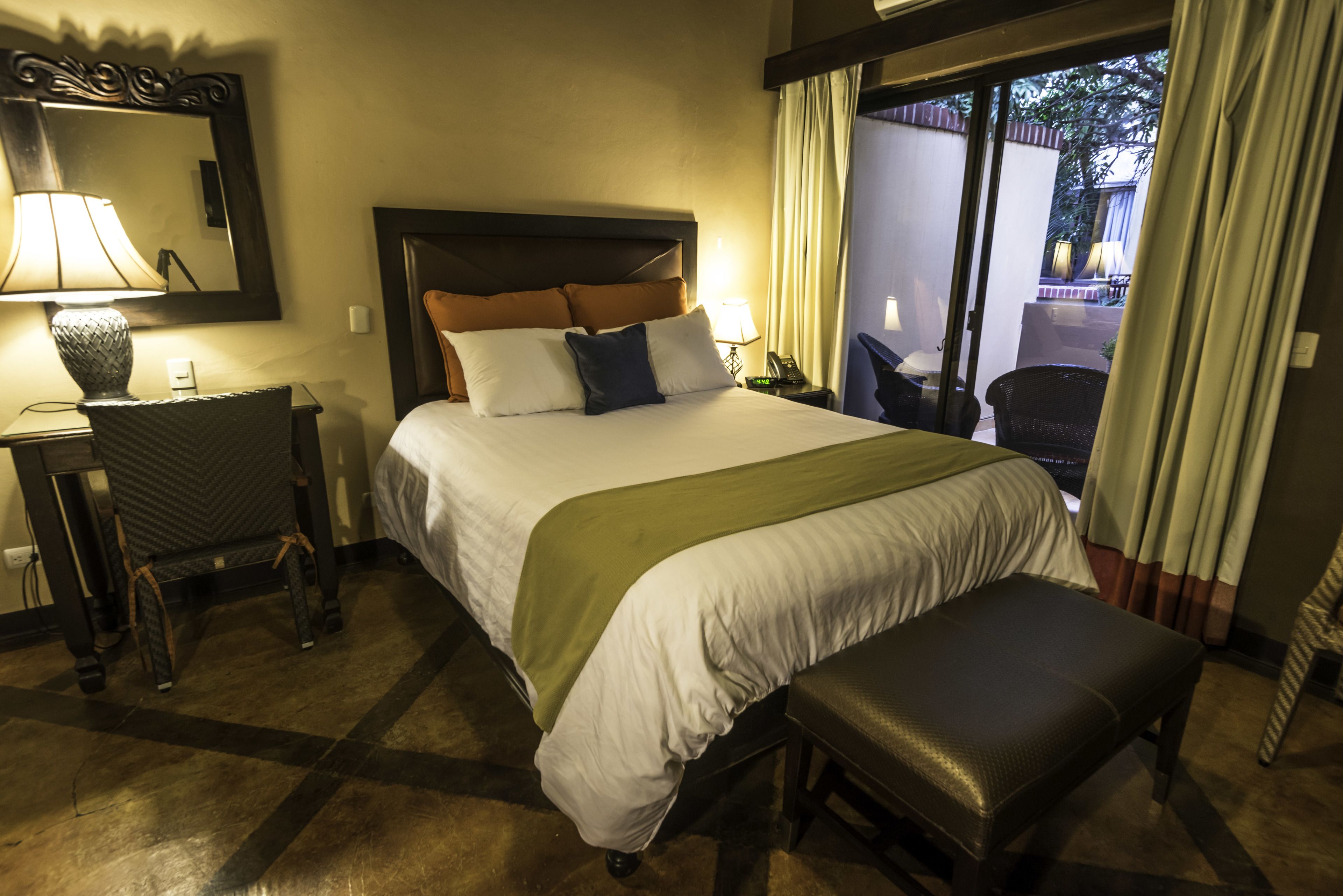 A hotel room with a large bed, a desk with a lamp, a chair, and a view of the outside through a window.