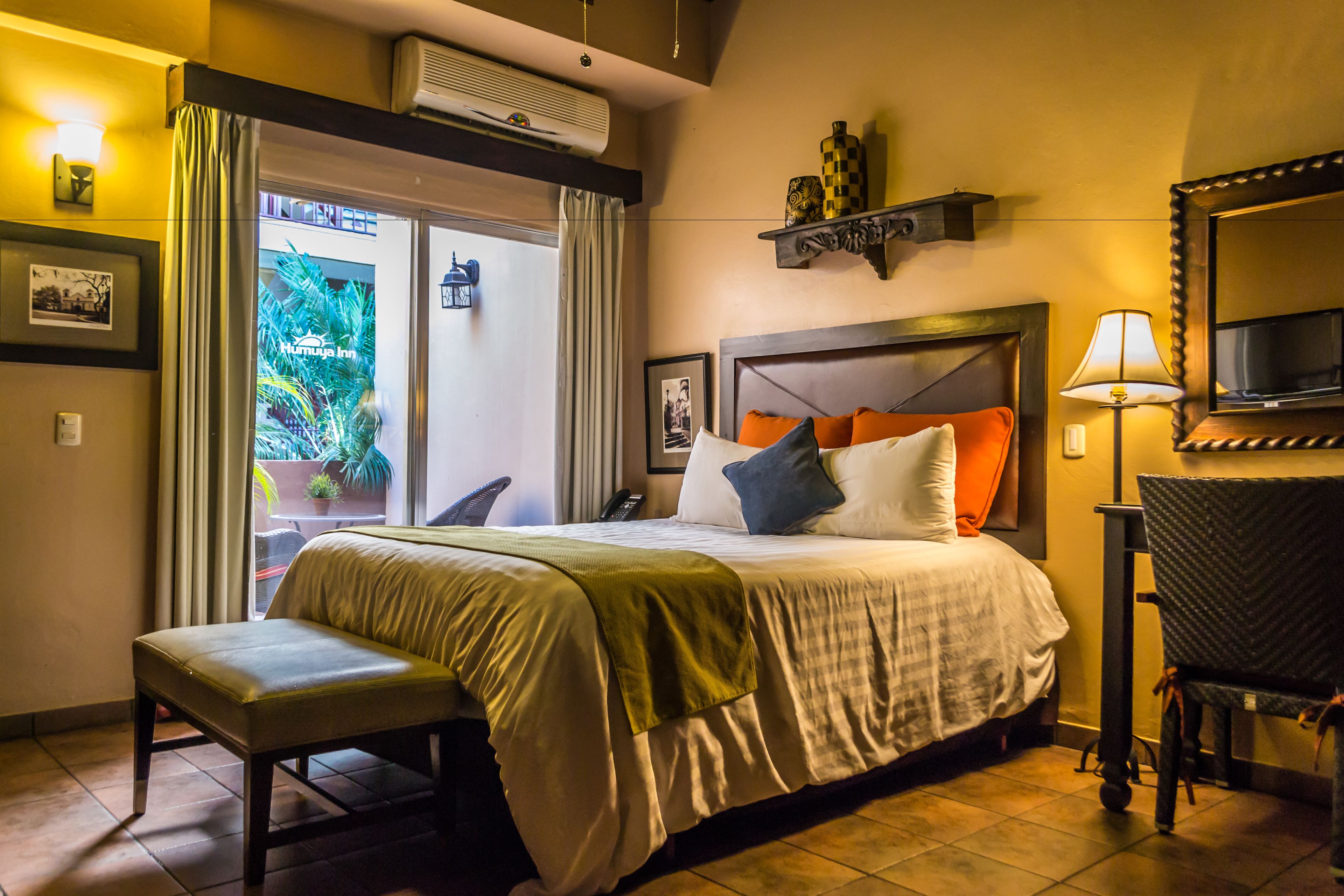 A hotel room with a bed, a bench, a desk, a TV, and a window with curtains.