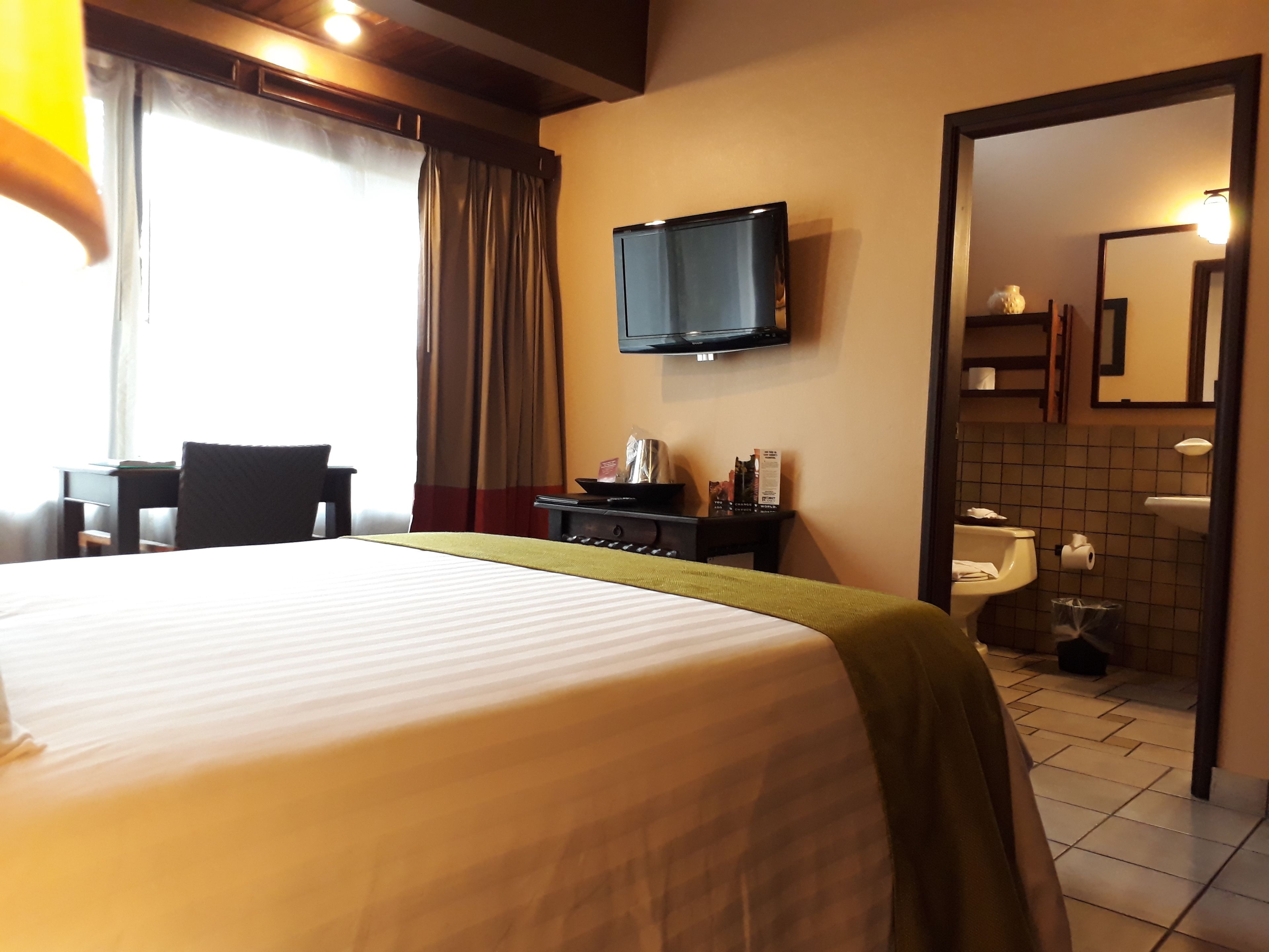Habitación de hotel con cama, escritorio, televisor montado en la pared y baño visible a través de una puerta abierta.