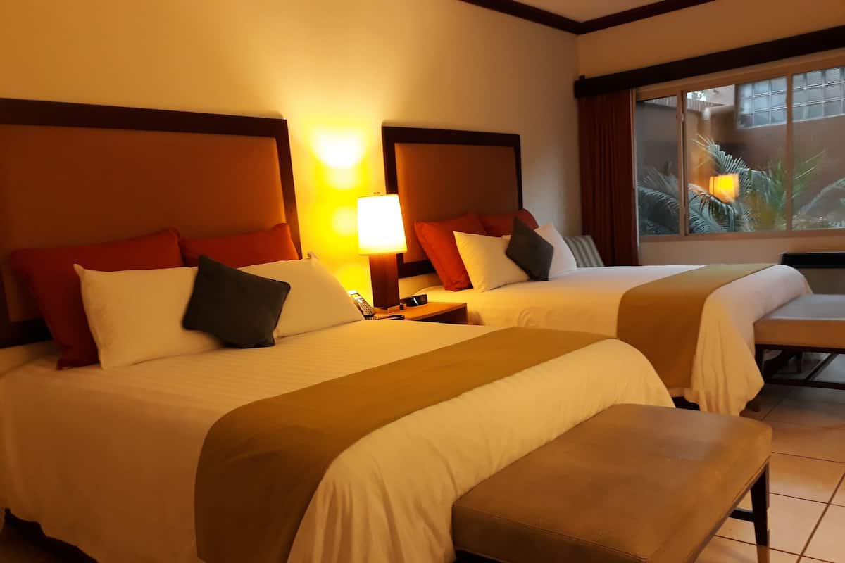 Una camera d'albergo con due letti, ognuno dotato di comodino, cuscini e lampada. Di fronte ai letti si trova una panca.