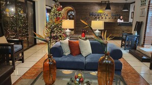 Um saguão de hotel com um sofá azul, dois vasos de vidro, um pinheiro de Natal e um balcão de recepção.