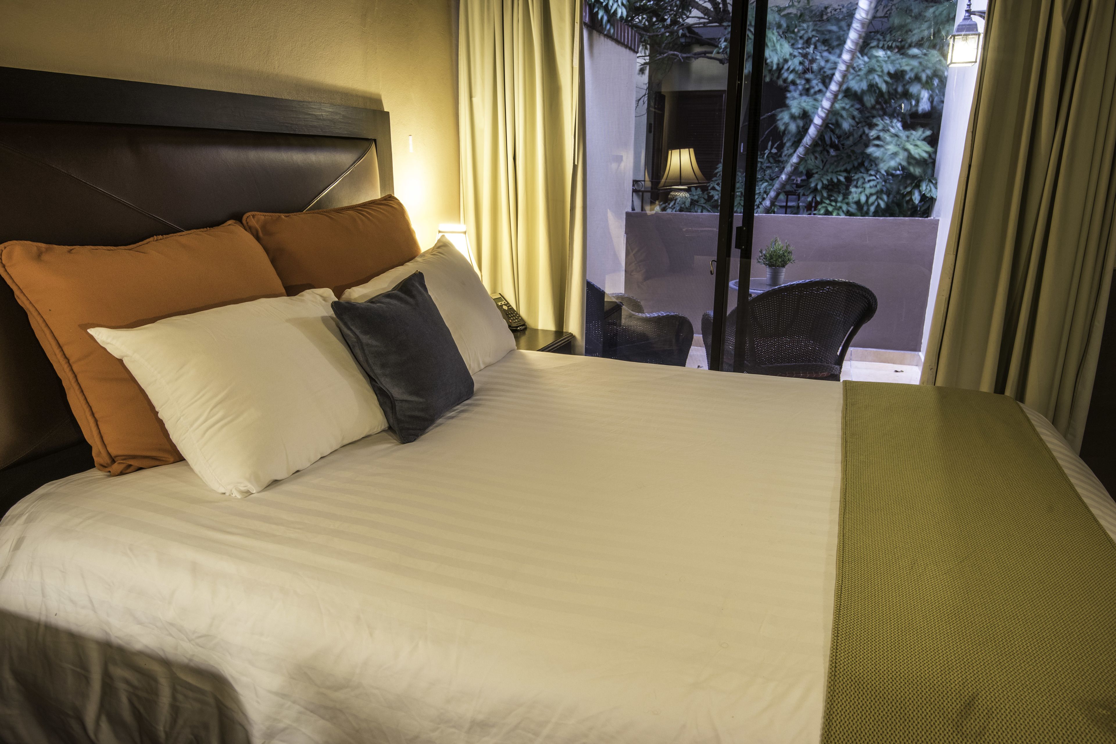 A hotel room with a large bed, a headboard, a bedside table with a lamp, and a view of the outside balcony.