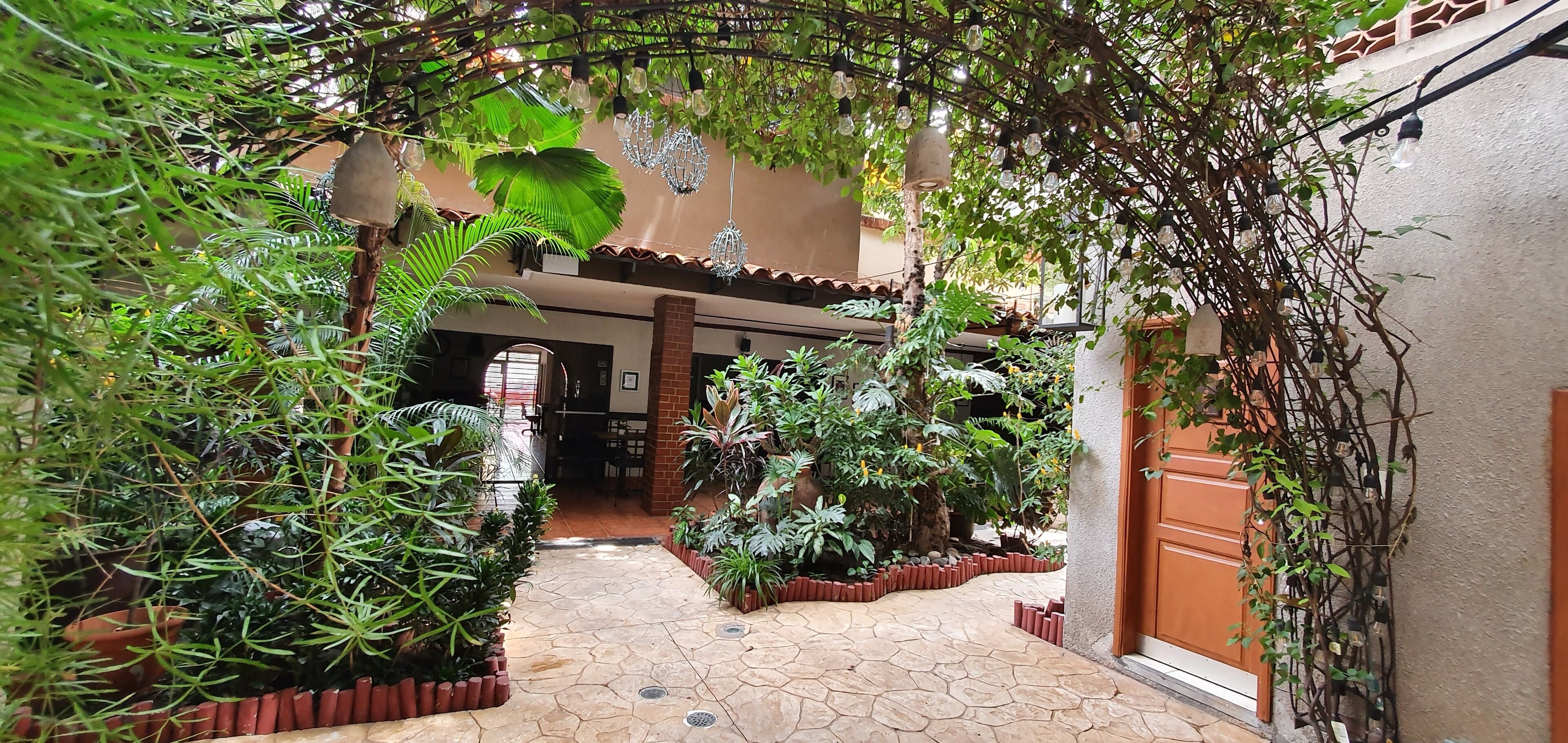 Un patio con sendero de piedra, plantas en macetas, lámparas colgantes y una puerta de madera.