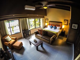 Habitación de hotel con cama, sofá, sillón, mesita de centro y escritorio. Cuenta con ventana con cortinas, ventilador de techo y unidad de aire acondicionado.