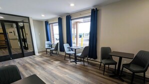 Et hotelværelse med trægulv, et spiseområde med stole og et bord, samt store vinduer med gardiner.