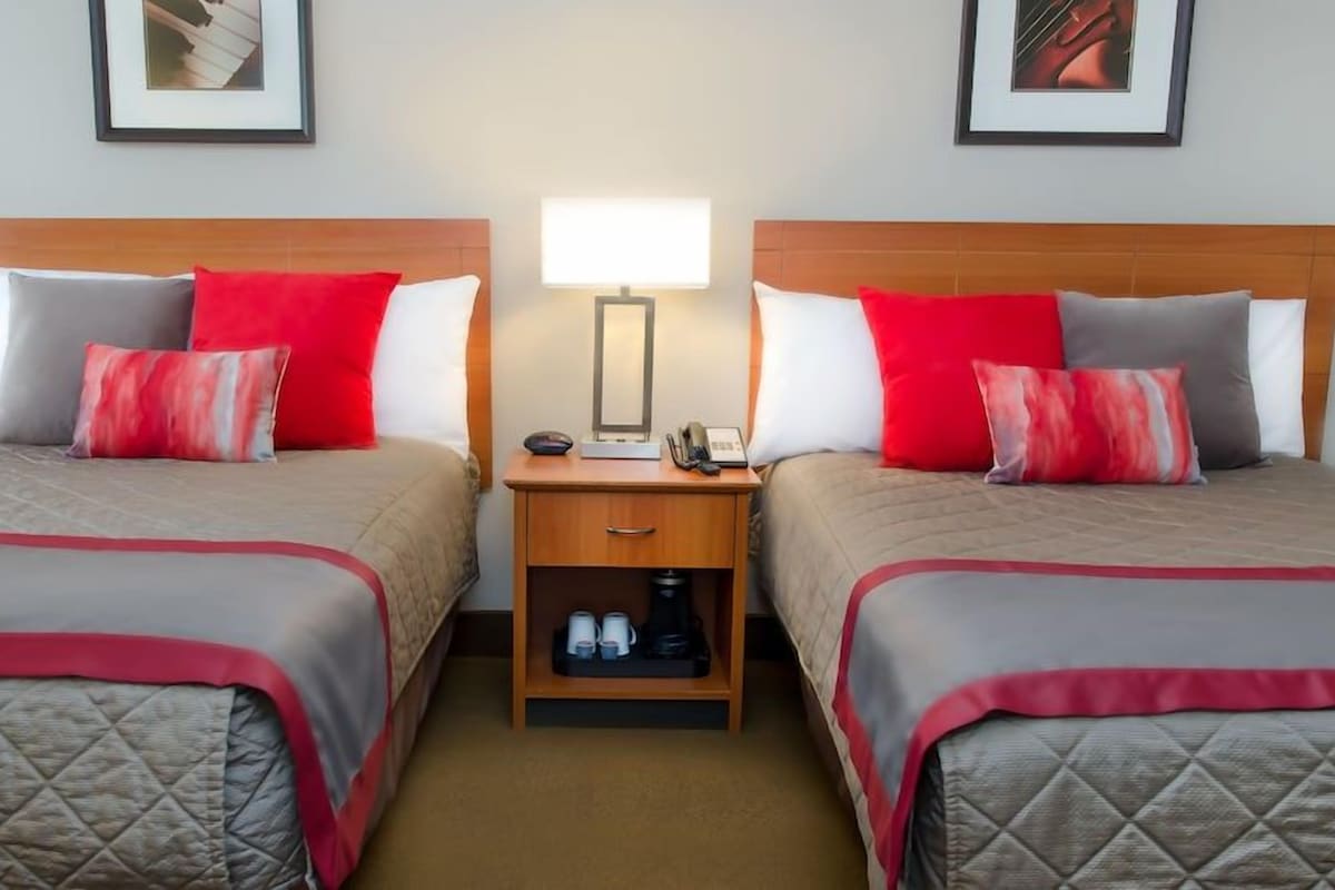 A hotel room with two beds, each with a red and white pillow, a bedside table with a lamp, and a small shelf with cups and a bottle.