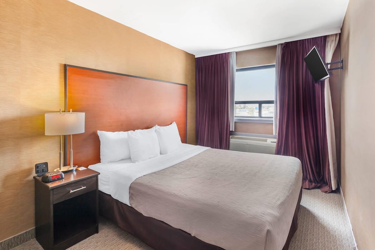 A hotel room with a large bed, a nightstand with a lamp, a TV mounted on the wall, and a window with curtains.