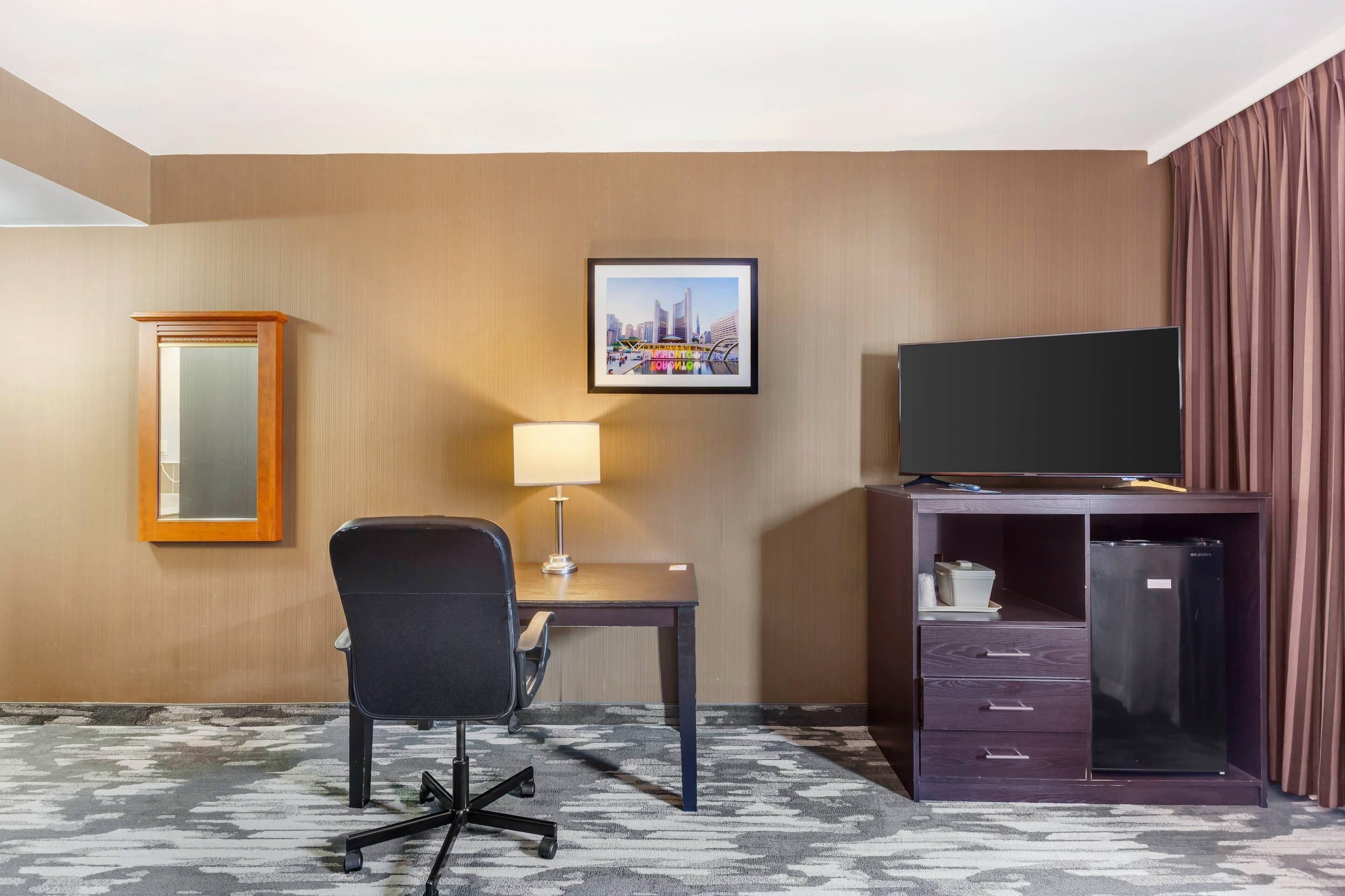 A hotel room with a desk, chair, television, minibar, and a framed picture on the wall.