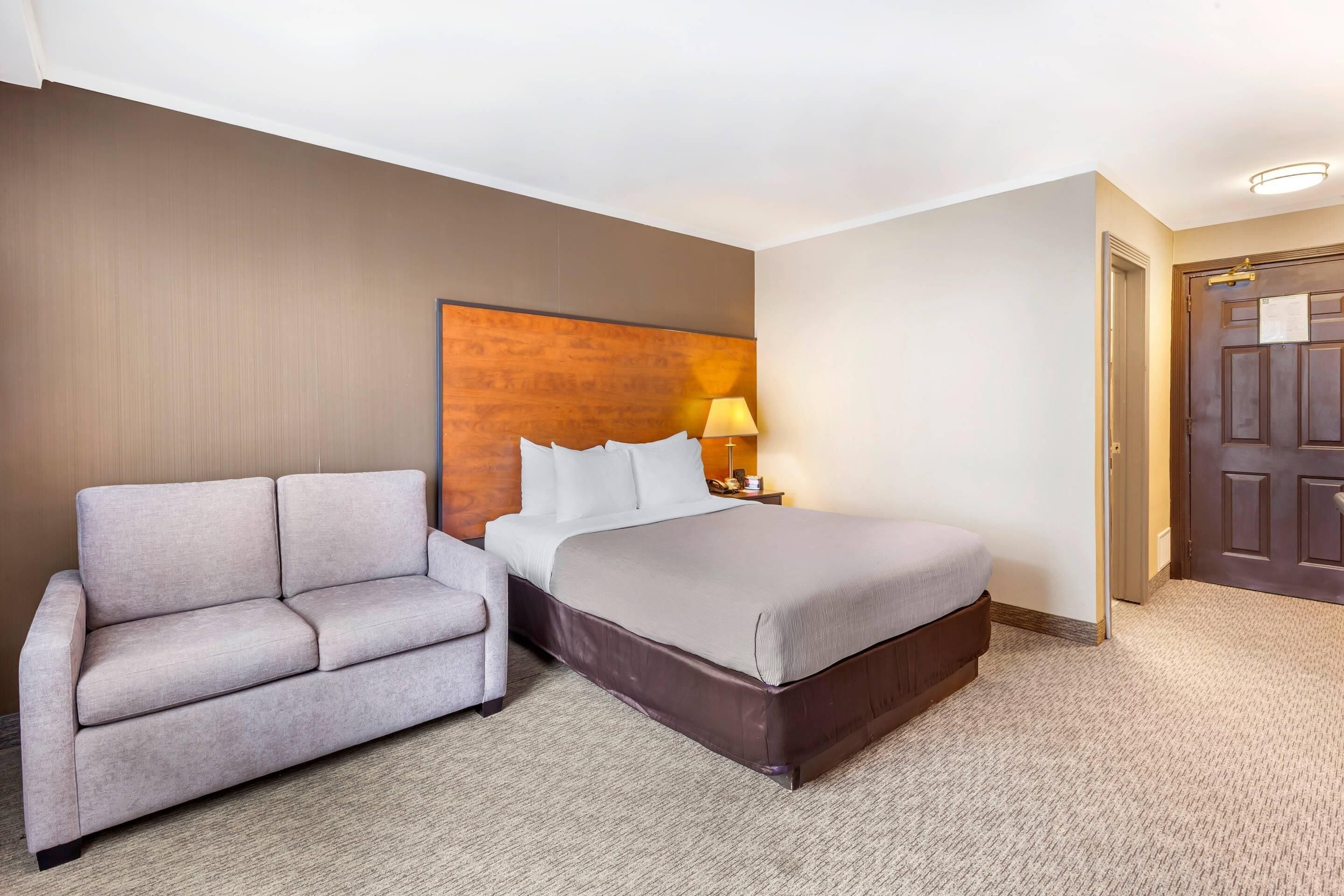 A hotel room with a bed, a gray sofa, a wooden headboard, a nightstand with a lamp, and a door.