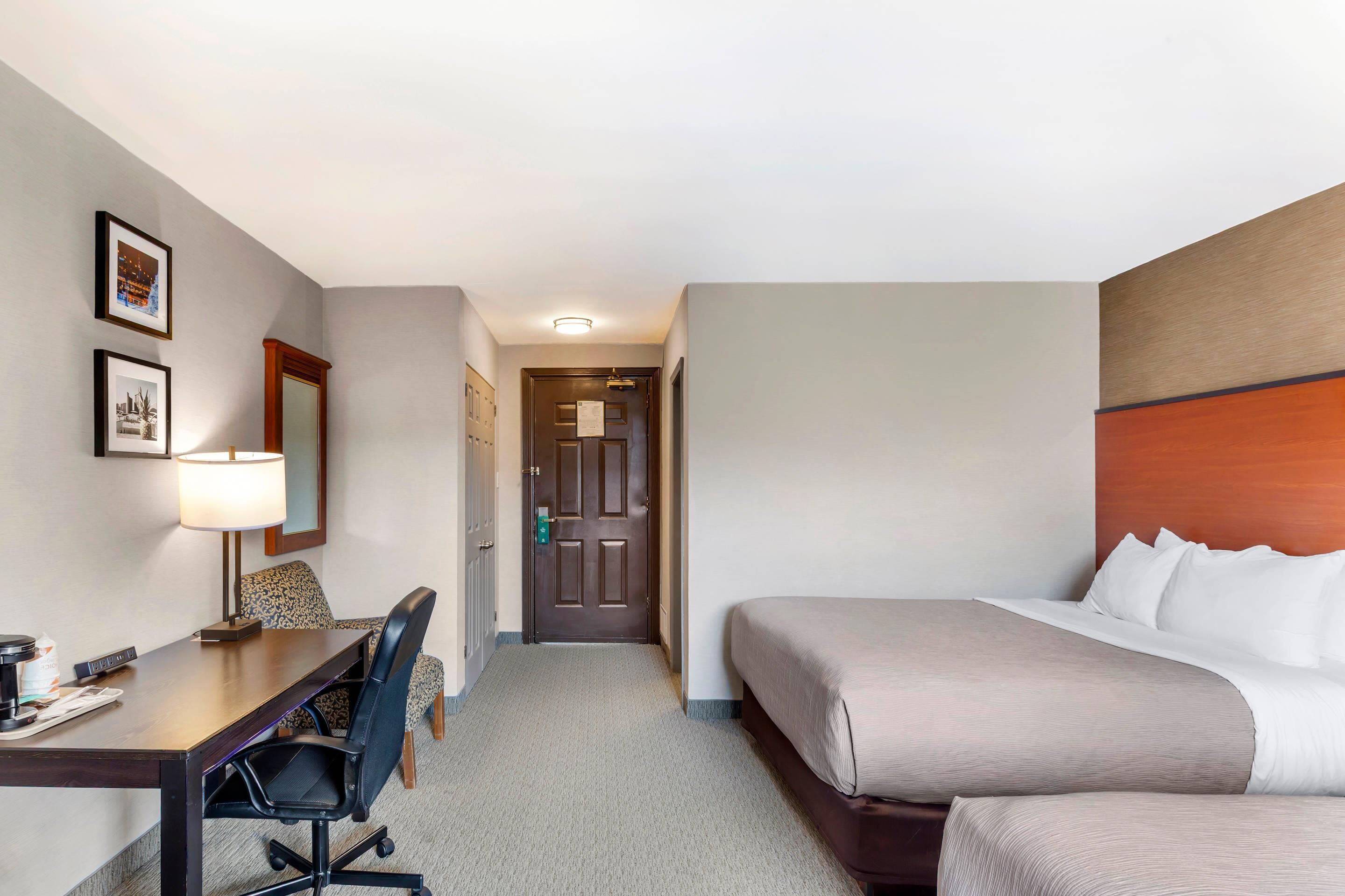 A hotel room with a bed, a desk with a chair, a lamp, and framed pictures on the wall.
