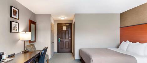 A hotel room with a bed, a desk with a chair, a lamp, and framed pictures on the wall.