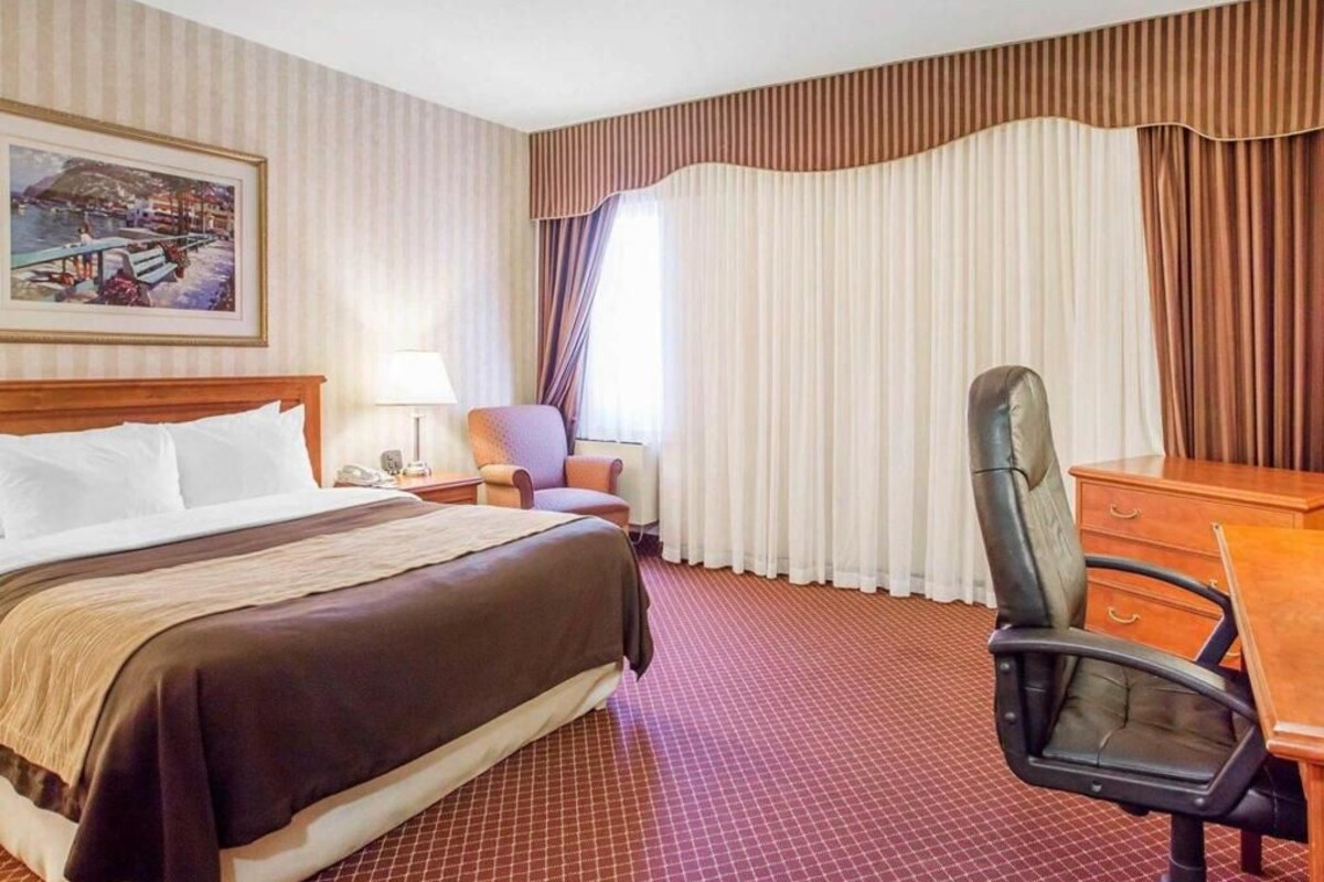 A hotel room with a bed, a desk, a chair, a lamp, and a painting on the wall.