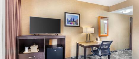 A hotel room with a flat-screen TV, a desk with a chair, a small refrigerator, a lamp, and a framed picture on the wall.