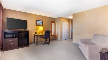 A hotel room with a flat-screen TV, a refrigerator, a desk with a lamp, a chair, and a sofa.